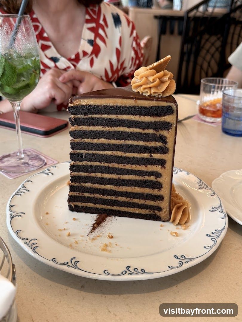 A tall, multi-layered chocolate cake with alternating layers of dark chocolate cake and peanut butter frosting sits on a white plate with blue trim. The cake is topped with a swirl of peanut butter frosting and a small wafer. A glass of minty green beverage and other drinks are visible in the background, along with a person wearing a patterned shirt.