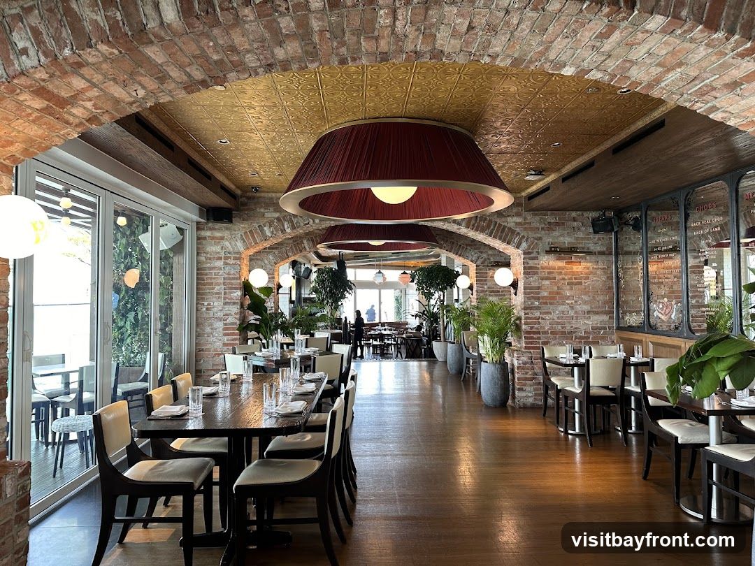 The interior of a restaurant with exposed brick walls, arched doorways, and a wooden floor. Large pendant lights hang from the ceiling. Tables are set with white tablecloths and chairs. Plants are placed throughout the space. In the background, through an archway, more tables and patrons can be seen. A menu board is visible on the right wall.