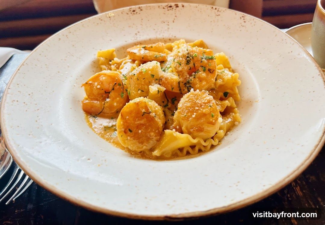 A close-up shot of a white plate filled with pasta, shrimp, and scallops in a creamy sauce, garnished with chopped herbs and grated cheese. The pasta appears to be a ruffled or wavy variety. The dish is presented on a dark, textured surface, with a hint of a napkin and cutlery visible on the left.