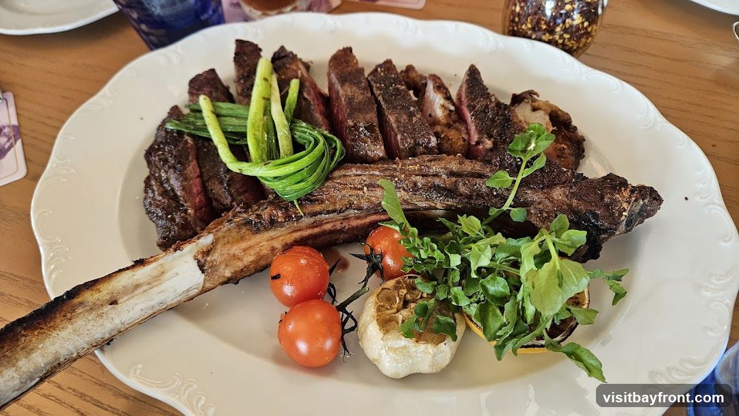 A large, sliced tomahawk steak is presented on a white platter with a decorative rim. The steak is cooked to a medium-rare, with a visible pink interior. It is garnished with grilled scallions, blistered cherry tomatoes, a head of roasted garlic, and a sprig of watercress. A lemon wedge is also visible. The platter sits on a wooden table. In the background, a small jar of chili flakes and a blue glass are partially visible.