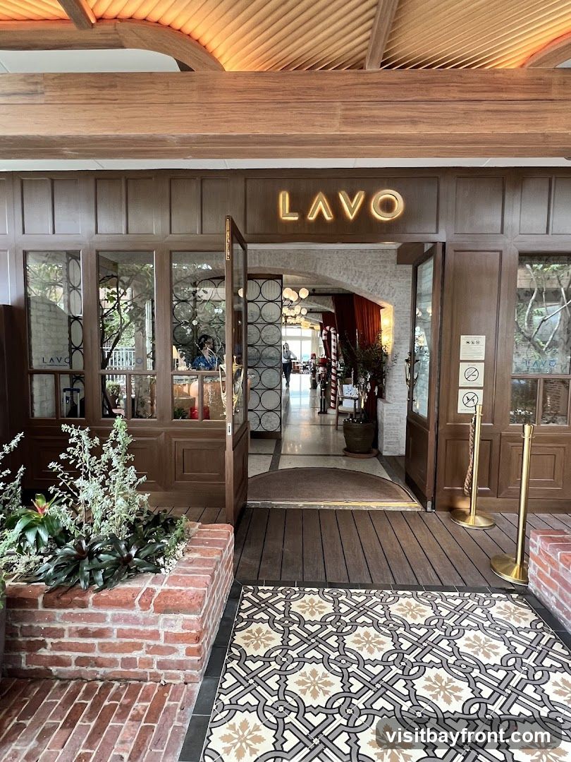The entrance to LAVO restaurant features a prominent illuminated sign above the doorway. The facade is constructed with wood paneling and glass windows. A patterned rug is laid out on the wooden walkway leading into the establishment.