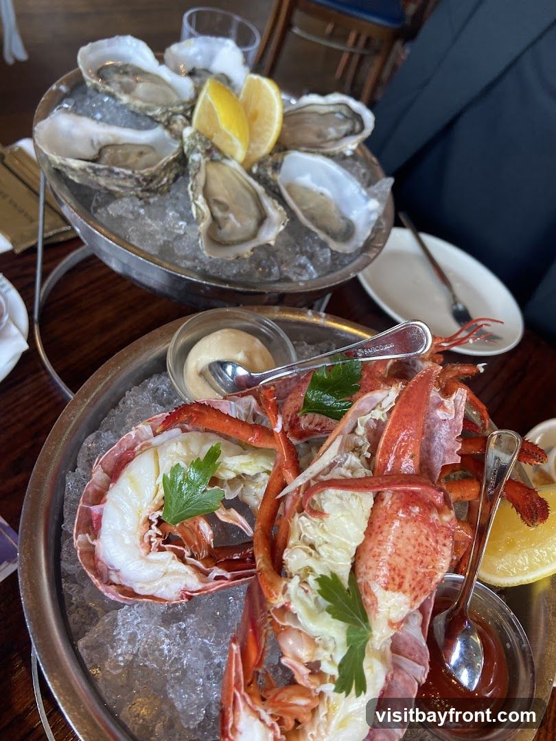 A close-up view of a seafood platter featuring a whole lobster and a selection of oysters on ice. The lobster is split in half, revealing its cooked meat, and garnished with parsley. The oysters are served on a tiered stand with lemon wedges. Small bowls of dipping sauces, likely cocktail sauce and a creamy sauce, are also visible.