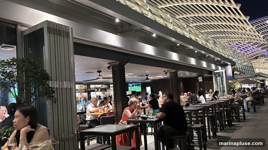 An outdoor dining area of a restaurant or bar at night. People are seated at tables, some inside the establishment and some on the patio. The interior features a bar with bottles on shelves and a television screen. The exterior has a modern, slatted roof structure overhead.