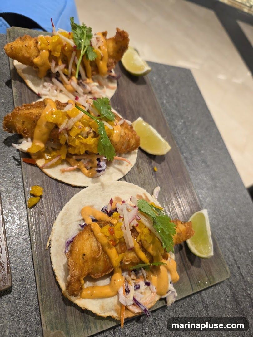 Three battered fish tacos are served on a dark slate platter. Each taco is filled with shredded cabbage, a creamy orange sauce, and topped with cilantro and a lime wedge.