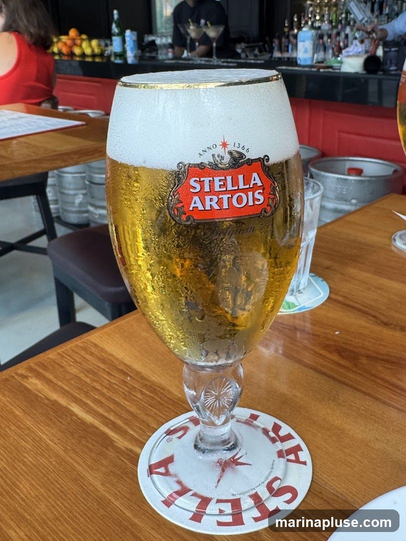 A close-up of a tall glass of Stella Artois beer, filled to the brim with golden liquid and topped with a thick layer of white foam. The iconic Stella Artois logo is prominently displayed on the glass. The glass sits on a wooden table, with a branded coaster beneath it. In the background, a bar area is visible with bottles and glasses, and a person wearing a red top is seated at a table.