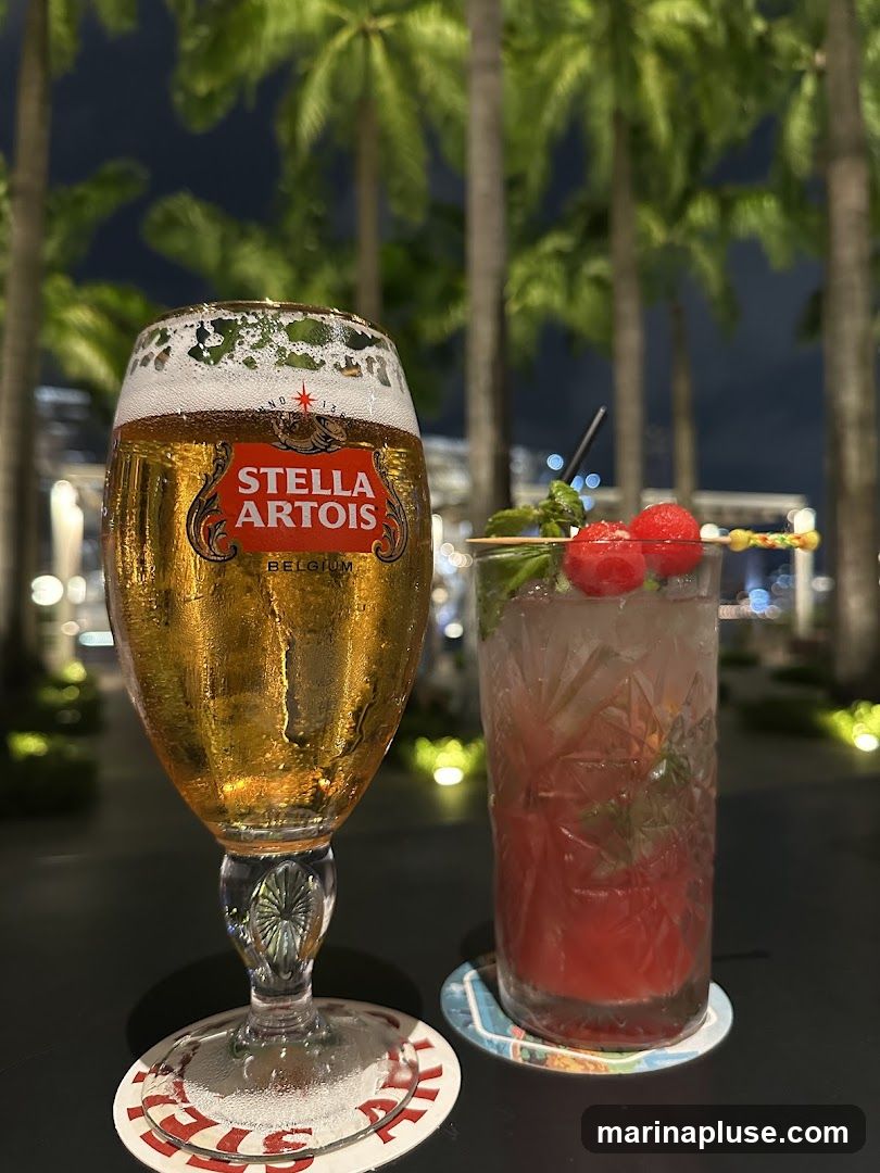 A glass of Stella Artois beer and a red cocktail with raspberries and mint are on a table at night, with palm trees and blurred lights in the background.
