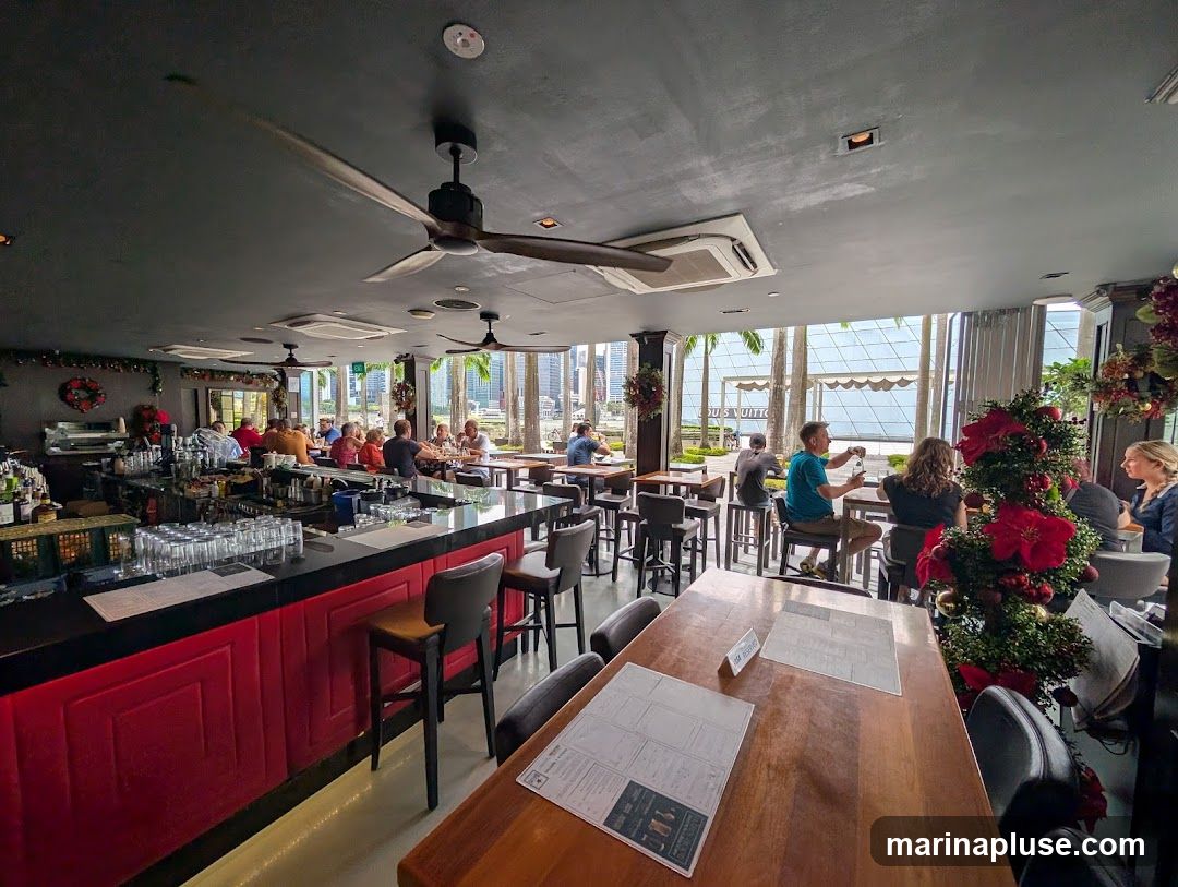 A view of a bar and restaurant interior with patrons seated at tables and the bar. The bar area is well-stocked with bottles and glassware. Large windows offer a view of palm trees and a city skyline. Christmas decorations, including wreaths and a decorated tree, are visible.