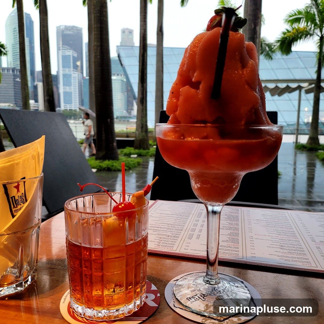 Two cocktails are on a wooden table, with a city skyline and palm trees in the background. One is a large frozen strawberry drink topped with a strawberry and a straw. The other is a darker amber-colored cocktail in a glass with a cherry and a pineapple wedge garnish. A menu is visible on the table.