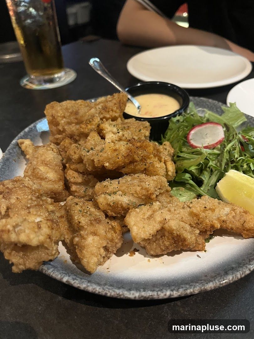 A plate of golden-brown fried chicken bites is served with a small side salad, a dipping sauce, and a lemon wedge. A glass of beer and plates are visible in the background.