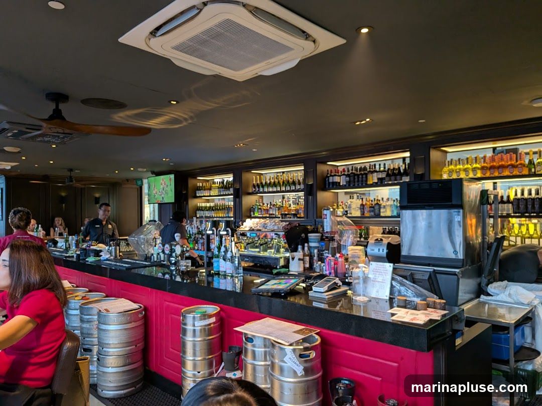 A busy bar interior with a long counter, shelves stocked with bottles of alcohol, and several staff members working. There are also kegs stacked near the counter and a television screen in the background.