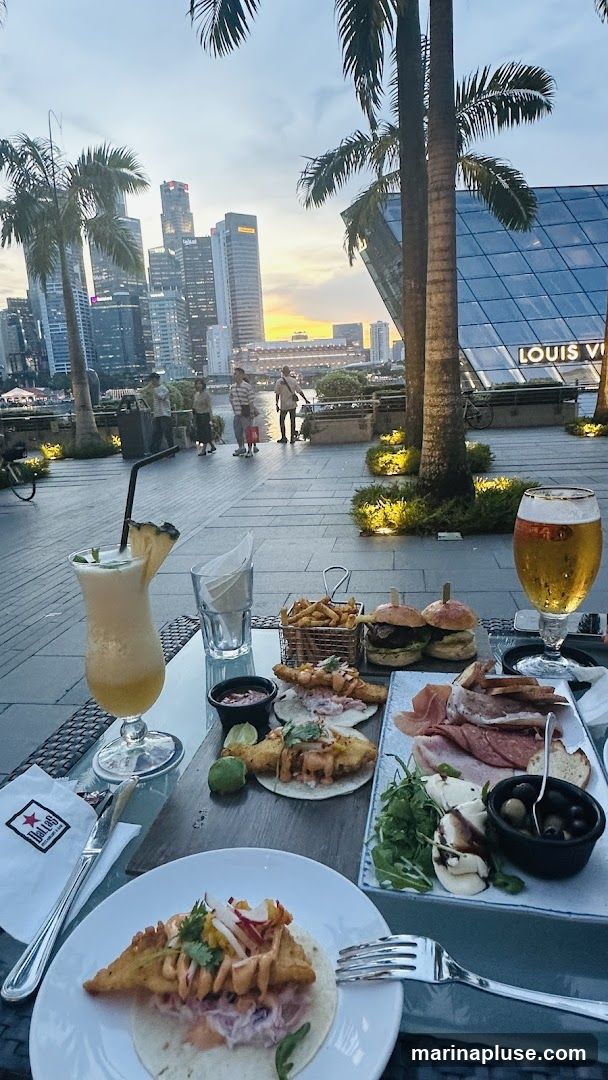 A table laden with food and drinks is set up outdoors, with a backdrop of a city skyline at dusk. On the table, there is a cocktail, a glass of beer, a platter of charcuterie and cheese, tacos, and sliders with fries. In the background, palm trees frame the view of modern skyscrapers and a body of water. The Louis Vuitton building is visible on the right. A napkin with the logo "Dallas Restaurant & Bar" is on the table.