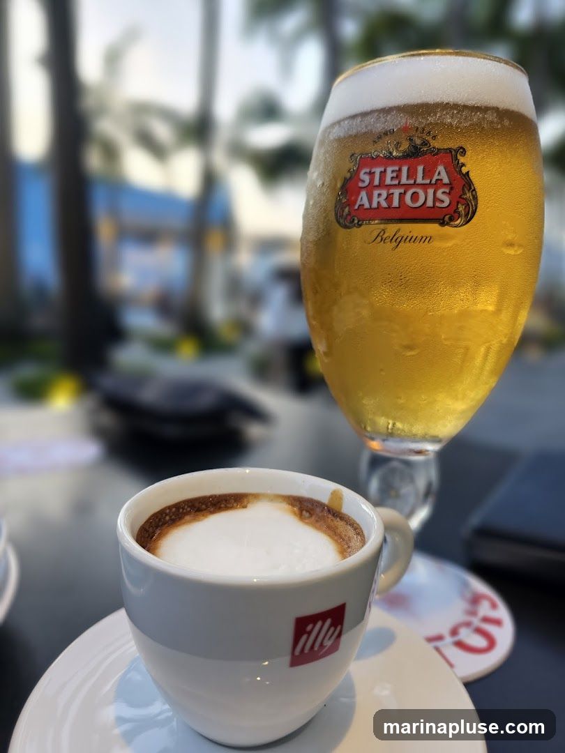 A close-up shot of a cappuccino in a white Illy cup and a glass of Stella Artois beer. The background is blurred, showing palm trees and a blue structure, suggesting an outdoor seating area at a cafe or bar.