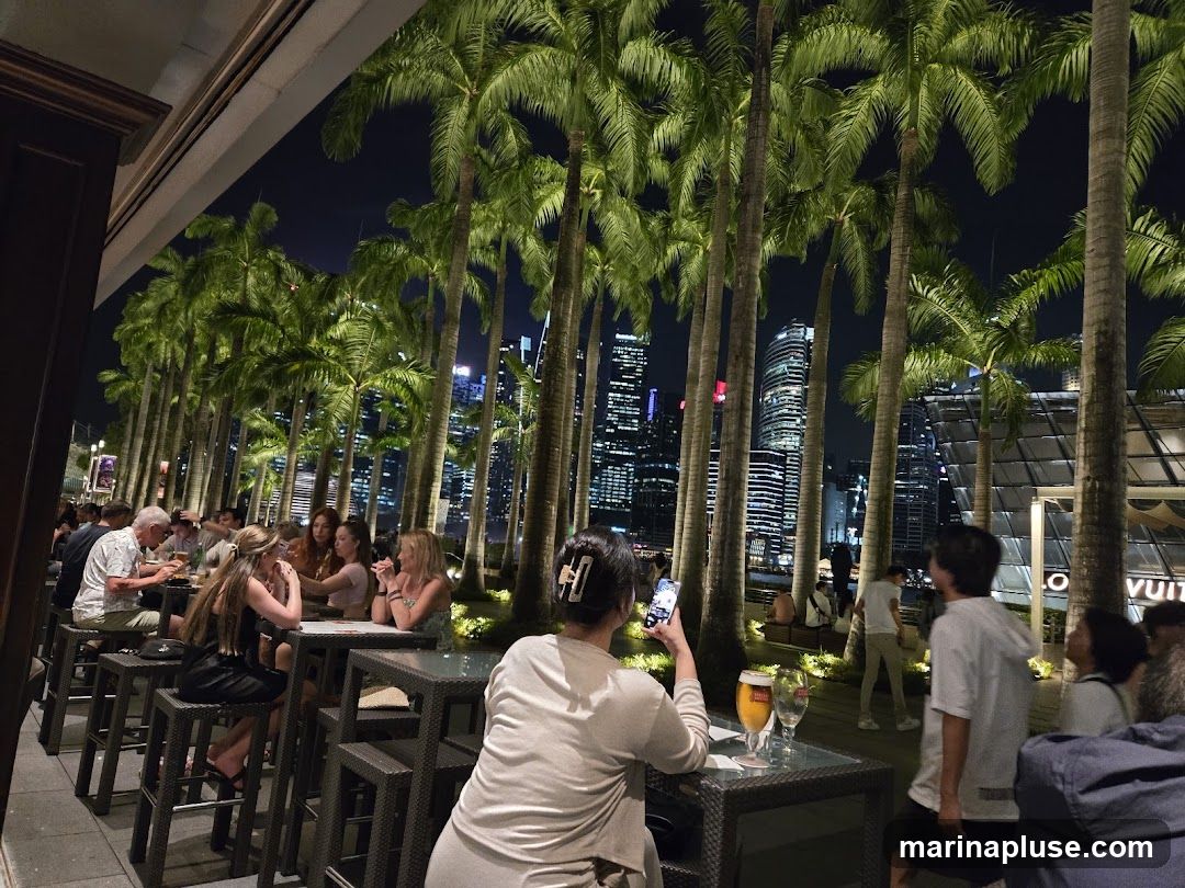 People dining at tables outdoors at night, with a backdrop of palm trees and a city skyline illuminated by lights. Some patrons are seated at high-top tables, while others are standing or walking. A Louis Vuitton sign is visible in the background.