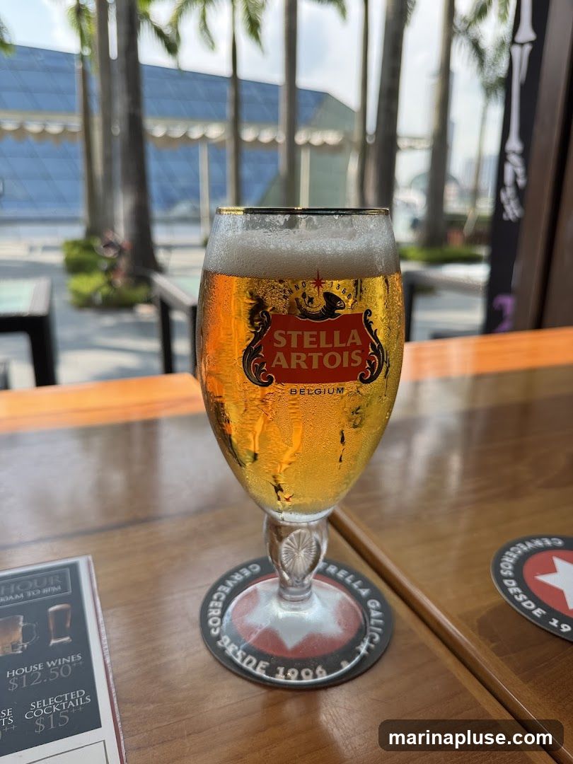 A glass of Stella Artois beer sits on a wooden table. In the background, palm trees and a modern building are visible. A coaster with "Estrella Galicia" is under the glass. A portion of a menu is visible on the left.