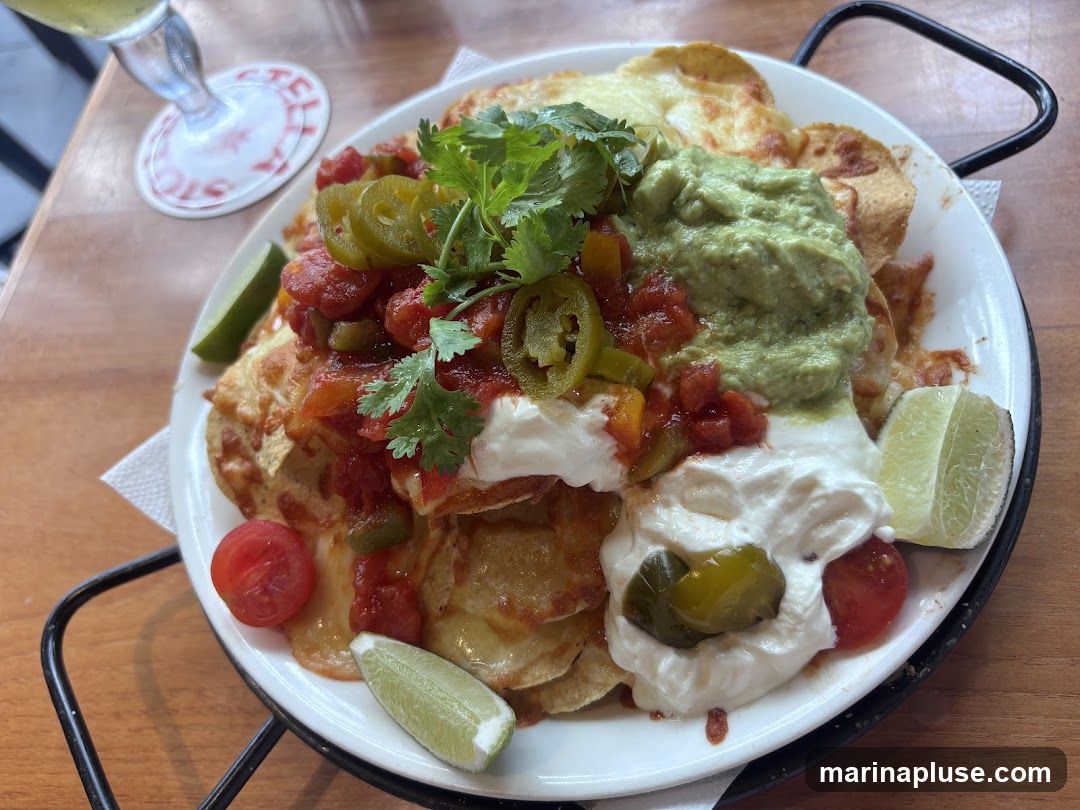 A plate of nachos topped with melted cheese, guacamole, salsa, sour cream, jalapenos, cilantro, cherry tomatoes, and lime wedges. The plate is served on a black metal stand on a wooden table. A coaster with red lettering is visible in the background.