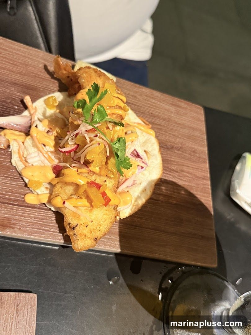 A close-up shot of a single fish taco served on a wooden board. The taco is filled with a piece of fried fish, shredded cabbage, red onion, and a drizzle of orange sauce, garnished with cilantro and diced yellow and red peppers.
