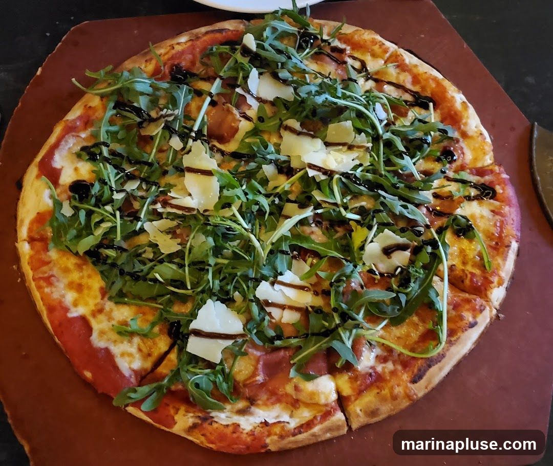 A close-up overhead view of a pizza served on a wooden board. The pizza has a golden crust, melted cheese, and is topped with fresh arugula, shaved parmesan cheese, and a drizzle of balsamic glaze.