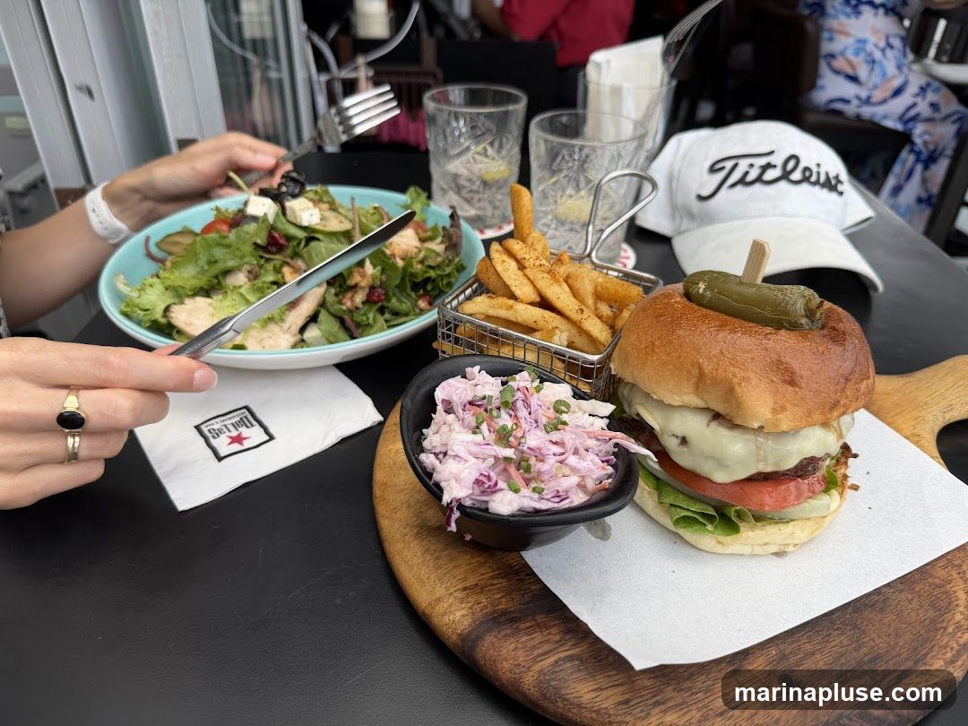 A close-up shot of a meal on a black table. In the foreground, a burger with melted cheese and a pickle on top is served with a side of coleslaw and a basket of french fries. To the left, a salad with chicken, feta cheese, and olives is being eaten by a person. A white baseball cap with the word "Titleist" is visible in the background.