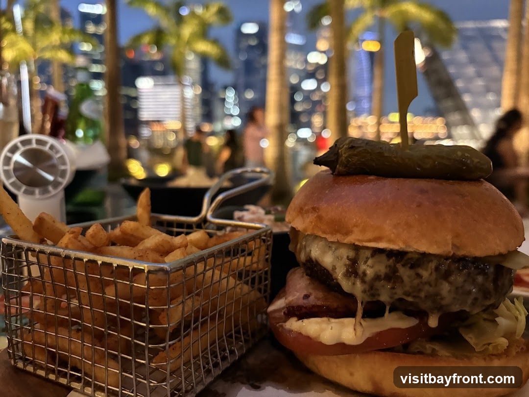 A close-up of a gourmet burger with a pickle on top and a side of french fries in a metal basket. The background shows a blurred cityscape at dusk with palm trees in the foreground, suggesting an outdoor dining setting.