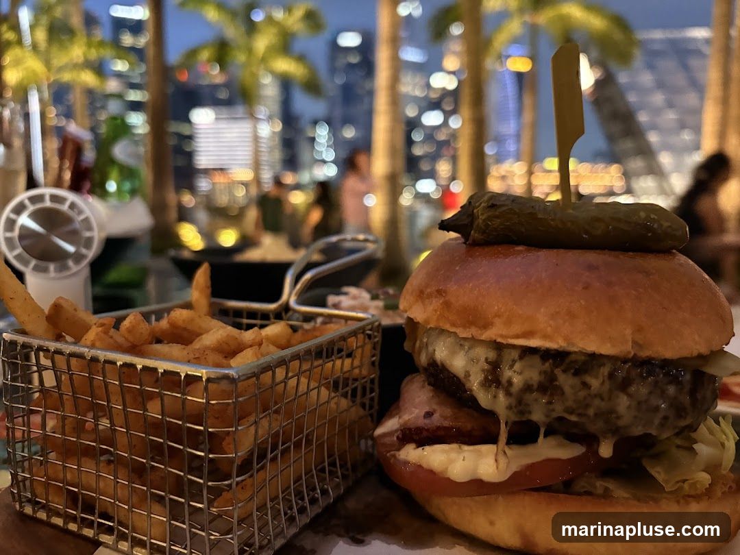 A close-up of a gourmet burger with a pickle on top and a side of french fries in a metal basket. The background shows a blurred cityscape at dusk with palm trees in the foreground, suggesting an outdoor dining setting.