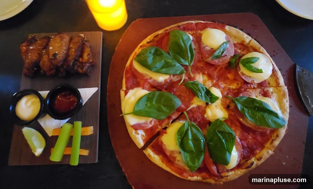 A close-up overhead shot of a pizza and chicken wings served on wooden boards. The pizza is a margherita style with fresh basil leaves, mozzarella, and tomato slices. The chicken wings are golden brown and seasoned with salt. Accompanying the wings are small bowls of dipping sauces, celery sticks, carrot sticks, and a lime wedge. A lit candle is visible in the background, casting a warm glow.
