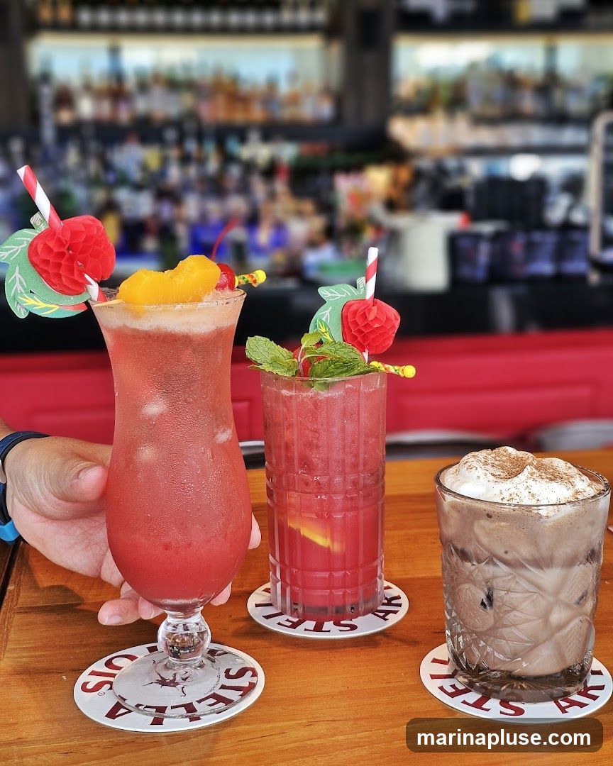 Three colorful cocktails are arranged on a wooden bar counter. The cocktail on the left is a tall hurricane glass filled with a red, icy beverage, garnished with a slice of orange and a cherry. The middle cocktail is in a fluted glass, also red, with mint leaves and a lemon slice. The glass on the right contains a chocolate-colored drink topped with whipped cream and a dusting of cinnamon. In the background, a blurred bar with shelves of bottles is visible. The coasters under the glasses have the word "STELLA" printed on them.