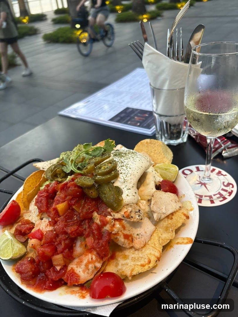 A plate of loaded nachos with chicken, salsa, jalapenos, and sour cream is served with a glass of white wine on a table outdoors. In the background, people are walking and cycling. A menu is visible on the table.