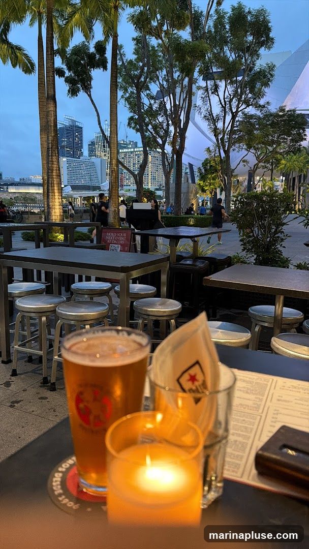 An outdoor seating area at dusk, with tables and stools set up. In the foreground, a glass of amber beer, a lit candle in a glass holder, and a menu are visible on a table. In the background, palm trees and other trees are silhouetted against a twilight sky, with city buildings and a modern architectural structure in the distance.