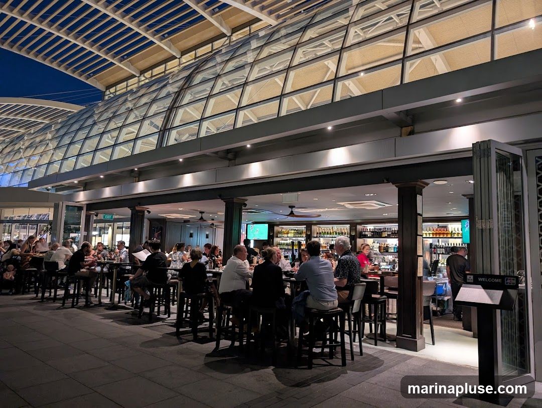 The image shows a lively bar and restaurant with many patrons seated at high-top tables and at the bar. The interior is well-lit, with shelves stocked with bottles behind the bar. A large, modern glass and metal roof structure extends over the outdoor seating area. The overall atmosphere appears to be social and bustling.