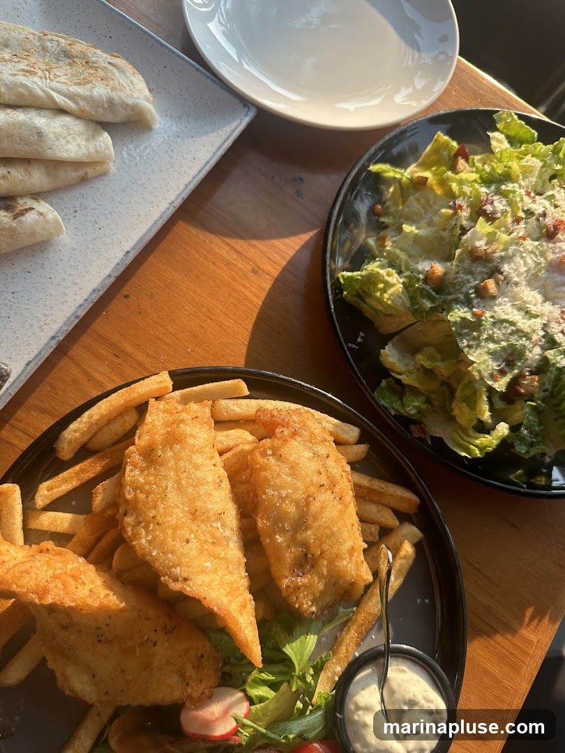 A close-up overhead shot of a meal on a wooden table. In the foreground, a plate of fish and chips is visible, with three pieces of battered fish served alongside a generous portion of french fries. A small dish of dipping sauce and a garnish of salad greens and radishes are also present. To the right, a black bowl contains a Caesar salad with croutons and grated cheese. In the upper left, a white rectangular plate holds several folded wraps or quesadillas. A white plate is stacked on top of another white plate in the background.