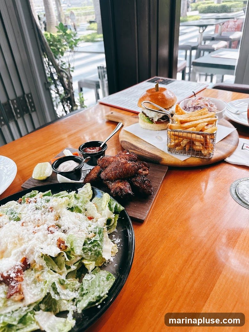 A table at a restaurant with a meal consisting of a burger, french fries, chicken wings, and a Caesar salad. The meal is served on a wooden table with a view of the outdoors through a window.