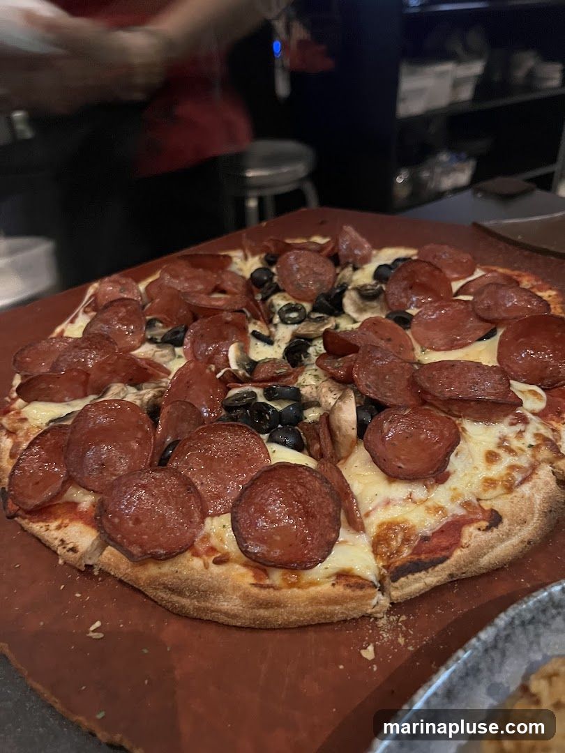 A close-up shot of a freshly baked pizza topped with generous amounts of pepperoni, sliced black olives, and mushrooms. The pizza has a golden-brown crust and is served on a wooden peel. In the background, a person in a red shirt is visible, along with some restaurant equipment.