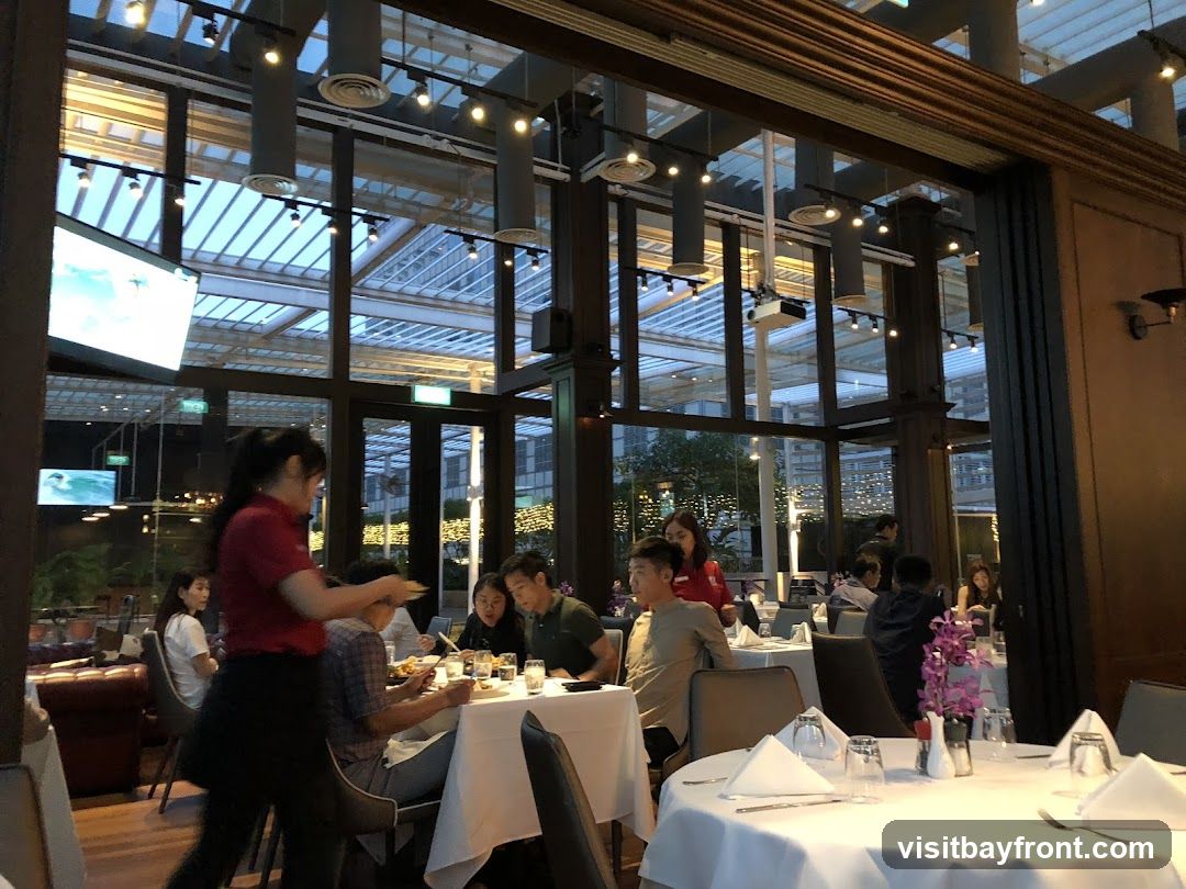 A busy restaurant interior with diners seated at tables and a server attending to a table. The restaurant has a modern design with a glass ceiling and large windows offering views of the outside.