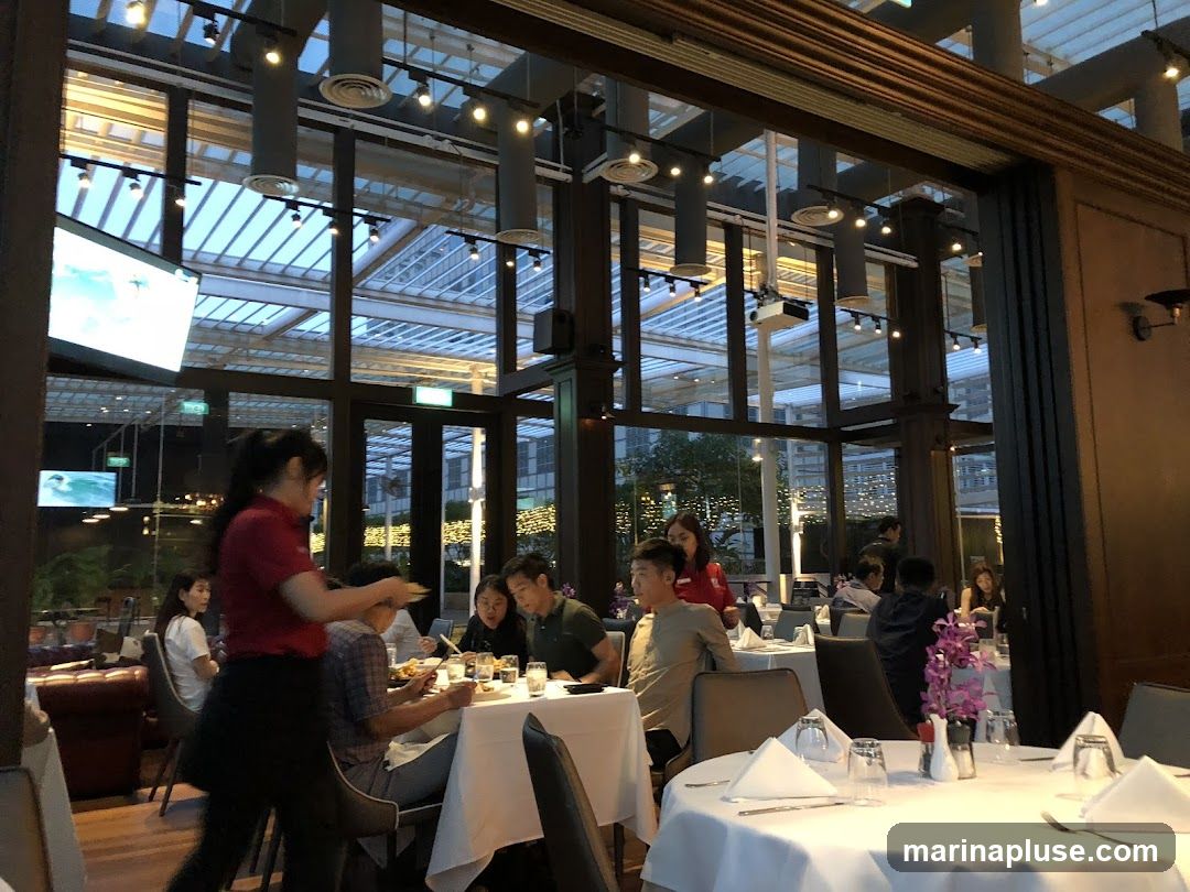 A busy restaurant interior with diners seated at tables and a server attending to a table. The restaurant has a modern design with a glass ceiling and large windows offering views of the outside.