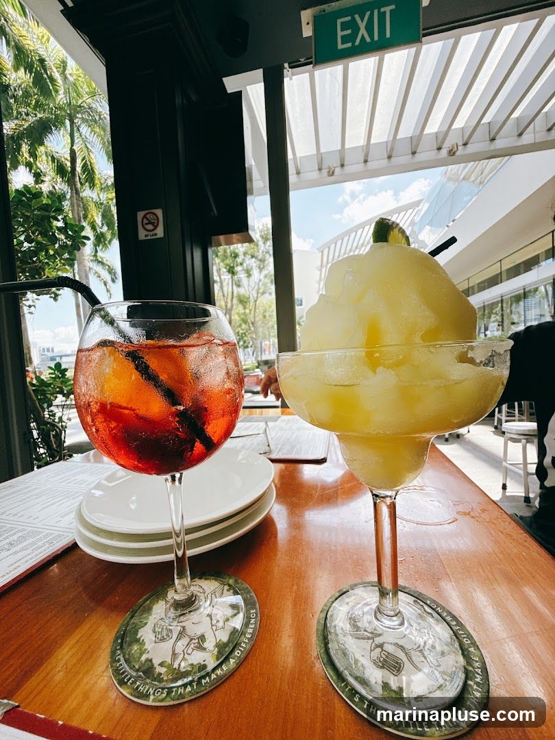 A close-up shot of two drinks on a wooden table. The drink on the left is a red cocktail with ice and a straw, served in a wine glass. The drink on the right is a yellow frozen drink with a lime wedge and a straw, served in a margarita glass. Both drinks have coasters underneath them with an illustration and text that reads "IT'S THE LITTLE THINGS THAT MAKE A DIFFERENCE". In the background, an "EXIT" sign is visible, along with palm trees and a bright sky through a window.