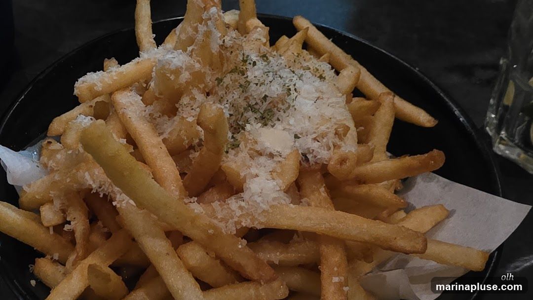 A close-up shot of a bowl of french fries, generously topped with grated parmesan cheese and herbs. The fries are golden brown and appear crispy. A small dollop of sauce is visible in the center. A glass with ice and lime wedges is partially visible in the background.