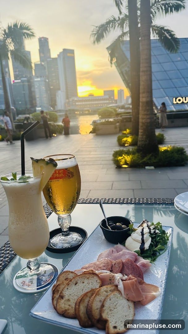 A table set for drinks and appetizers with a scenic view of a city skyline during sunset. On the table, there is a creamy cocktail with a pineapple slice and mint, a glass of beer, and a platter with assorted cured meats, bread slices, mozzarella cheese, arugula, and olives. In the background, palm trees frame the view of modern skyscrapers and a body of water reflecting the warm hues of the setting sun.