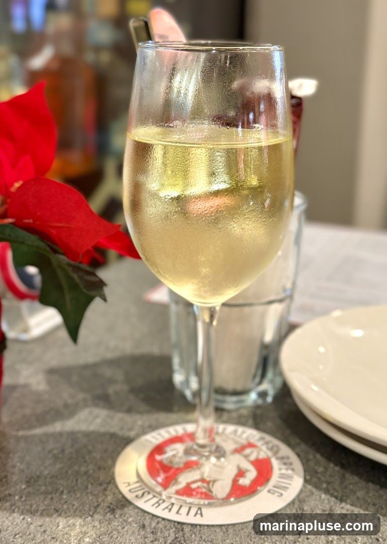 A close-up shot of a glass filled with white wine, condensation visible on the glass. The glass sits on a coaster with a red and white circular logo that reads "AUSTRALIA" and appears to depict a winged figure. In the background, a red poinsettia flower is visible, along with a blurred interior setting that includes bottles and plates.