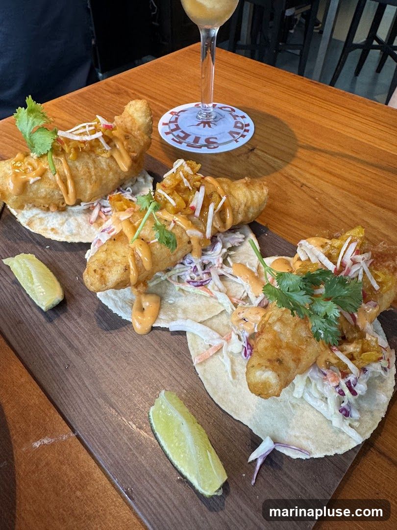Three fish tacos served on a wooden board with lime wedges. The tacos are topped with slaw and a drizzle of sauce, and garnished with cilantro. A glass of beverage and a coaster with a logo are visible in the background.