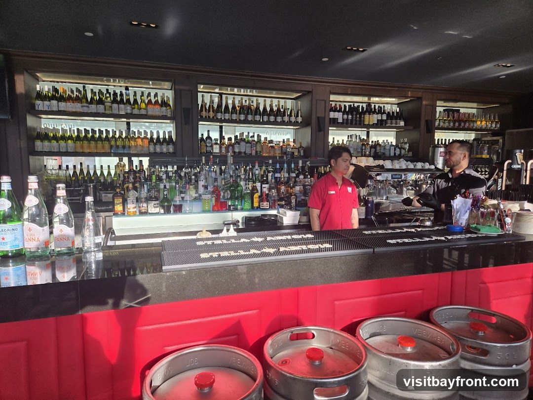 A well-stocked bar with bottles of wine and spirits displayed on shelves. Two bartenders are visible behind the counter, one in a red uniform and the other in a grey shirt. In the foreground, several beer kegs are stacked.