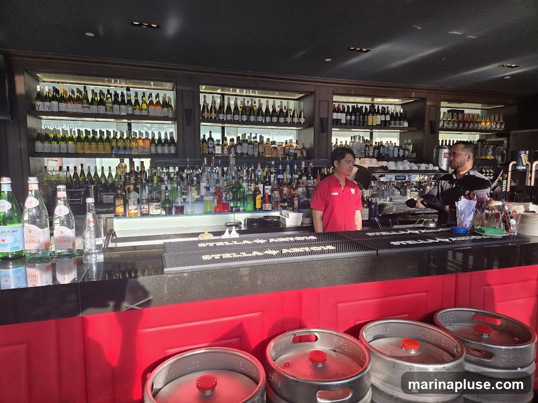 A well-stocked bar with bottles of wine and spirits displayed on shelves. Two bartenders are visible behind the counter, one in a red uniform and the other in a grey shirt. In the foreground, several beer kegs are stacked.