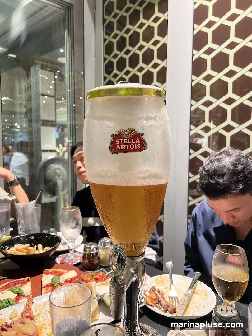 A table at a restaurant is set with a large beer tower featuring the Stella Artois logo. Various food items are visible, including pizza, fries, and what appears to be meat dishes. Several people are seated around the table, engaged in dining. The background shows decorative wall panels and reflections in a window.