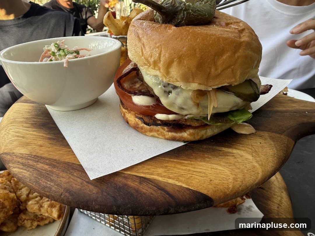 A close-up shot of a large burger served on a wooden board. The burger is topped with a pickled pepper and appears to have bacon, cheese, tomato, lettuce, and pickles. To the left of the burger is a white bowl filled with coleslaw. In the background, partially visible, are french fries and other fried food items. People are seated at the table, suggesting a dining setting.