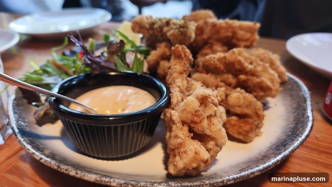 A plate of fried calamari served with a creamy dipping sauce and a small side salad. The calamari is golden brown and appears crispy. The dipping sauce is a light orange color with herbs. The salad consists of mixed greens. The plate is set on a wooden table.