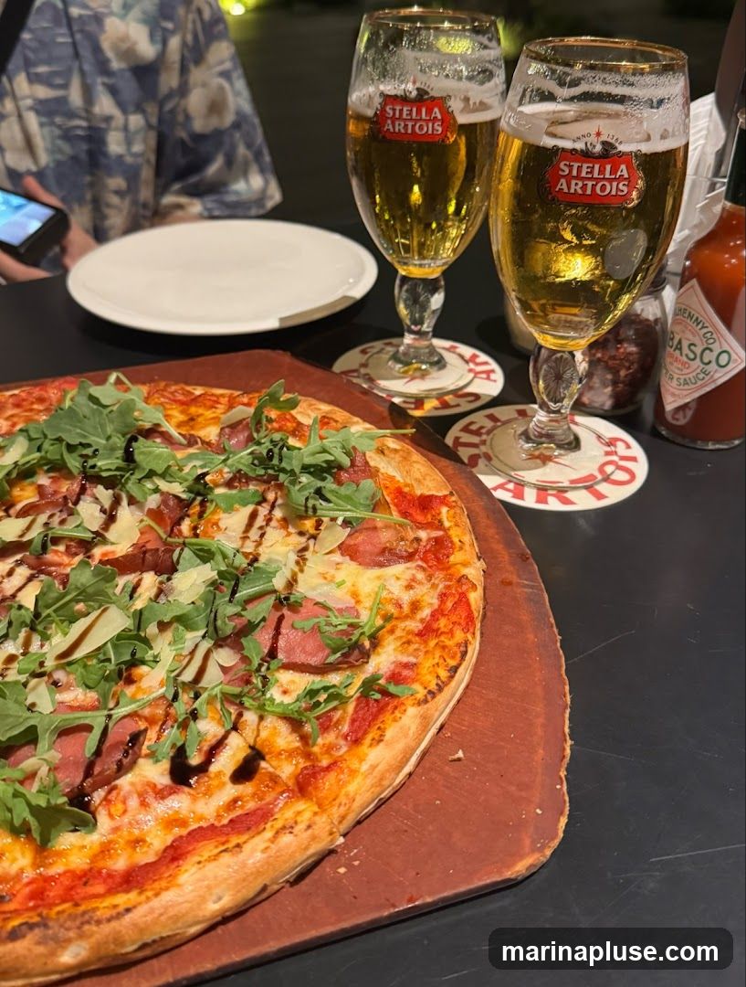 A close-up shot of a pizza and two glasses of Stella Artois beer on a dark table. The pizza is topped with arugula, meat, and cheese, and drizzled with balsamic glaze. A bottle of Tabasco sauce is visible in the background.