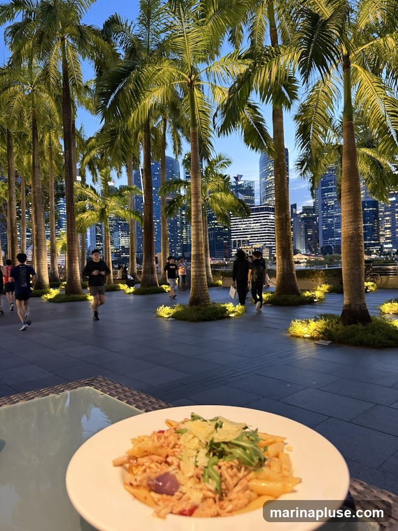 A plate of pasta with shredded meat, arugula, and parmesan cheese sits on a table in the foreground. In the background, a line of palm trees separates a paved walkway from a city skyline with illuminated skyscrapers. People are seen walking and jogging on the walkway. The scene is set at dusk.
