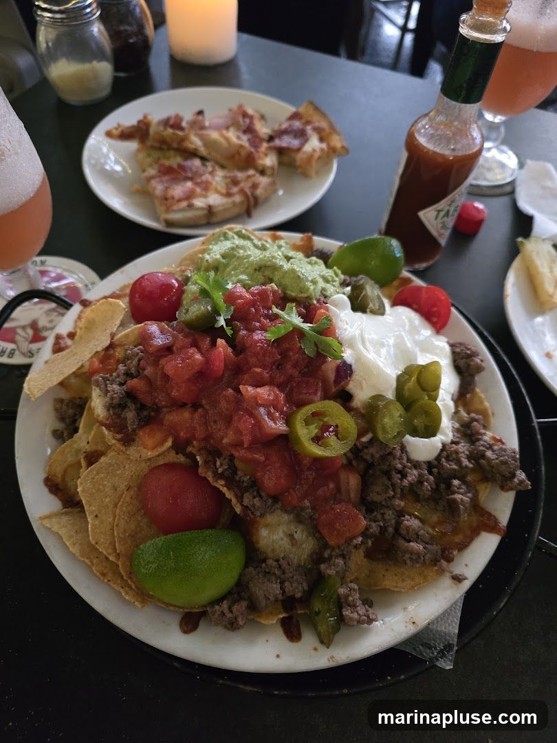 A plate of loaded nachos with ground beef, salsa, guacamole, sour cream, jalapenos, and tomatoes is in the foreground. Behind it, a plate of pizza slices is visible. A bottle of Tabasco sauce and a glass of pink beverage are also on the table. A lit candle is in the background.