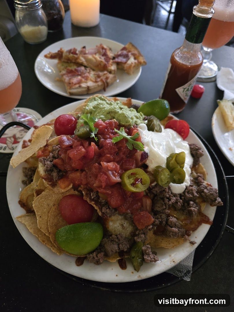 A plate of loaded nachos with ground beef, salsa, guacamole, sour cream, jalapenos, and tomatoes is in the foreground. Behind it, a plate of pizza slices is visible. A bottle of Tabasco sauce and a glass of pink beverage are also on the table. A lit candle is in the background.