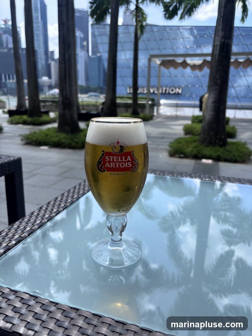 A glass of Stella Artois beer with a foamy head sits on a glass-topped table. In the background, palm trees and modern buildings are visible, including a building with the Louis Vuitton logo. The scene appears to be an outdoor seating area.