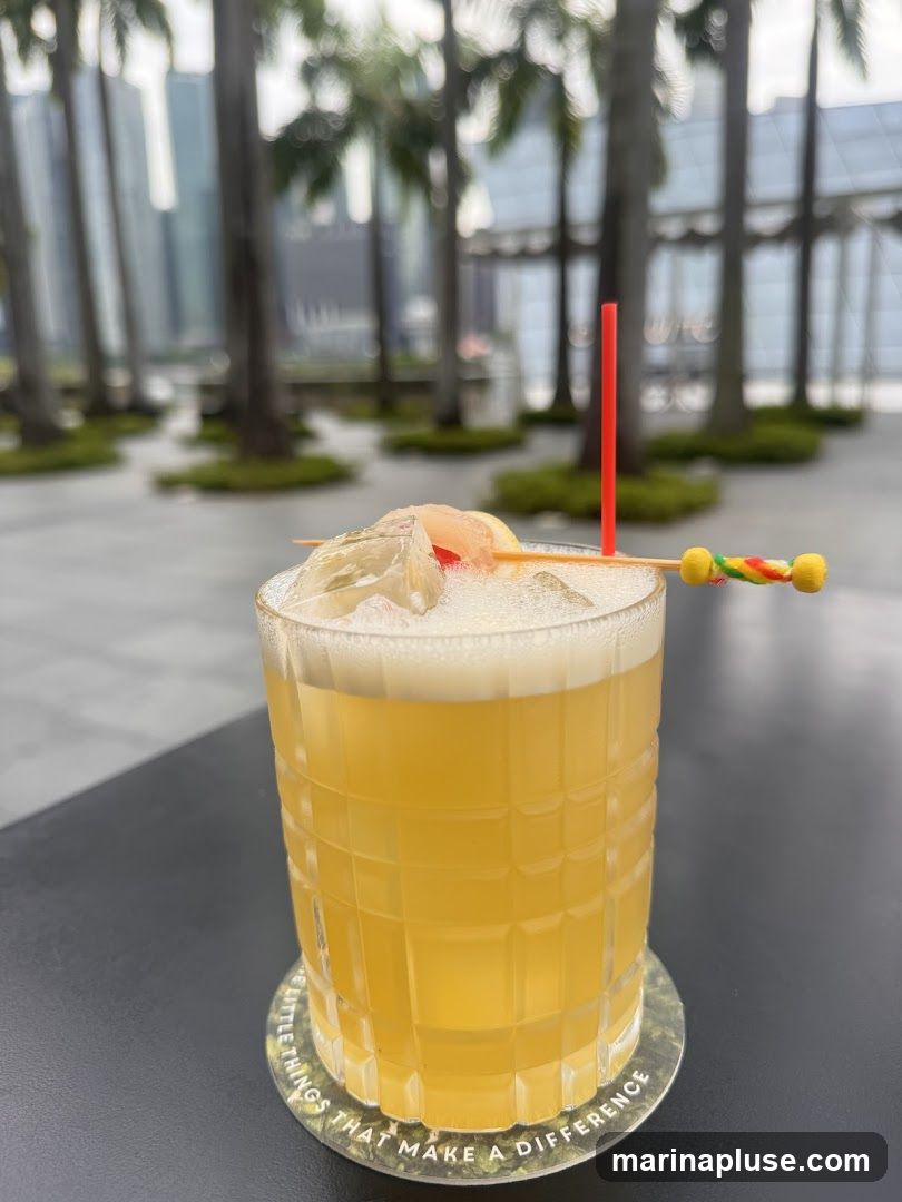 A close-up shot of a yellow cocktail in a textured glass, garnished with ice, a slice of fruit, and a red straw. The drink sits on a coaster with text that reads "LITTLE THINGS THAT MAKE A DIFFERENCE." In the blurred background, palm trees and modern buildings suggest an urban, tropical setting.